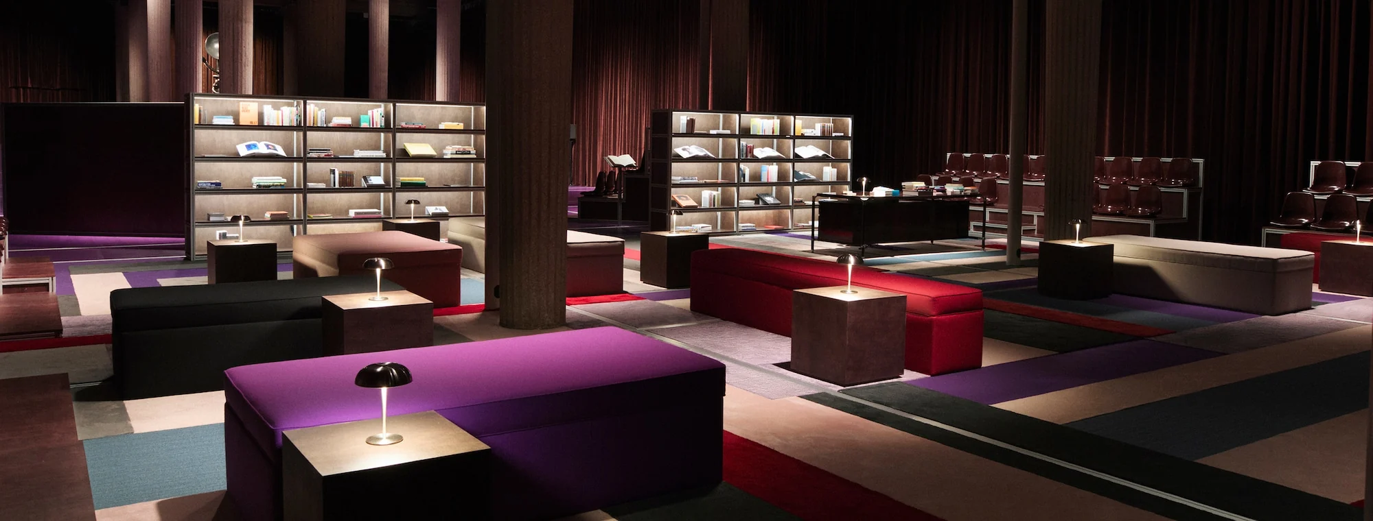 Illuminated bookshelves, upholstered benches and reading lamps form a library among concrete columns and dark curtains at the Palais d’Iéna.