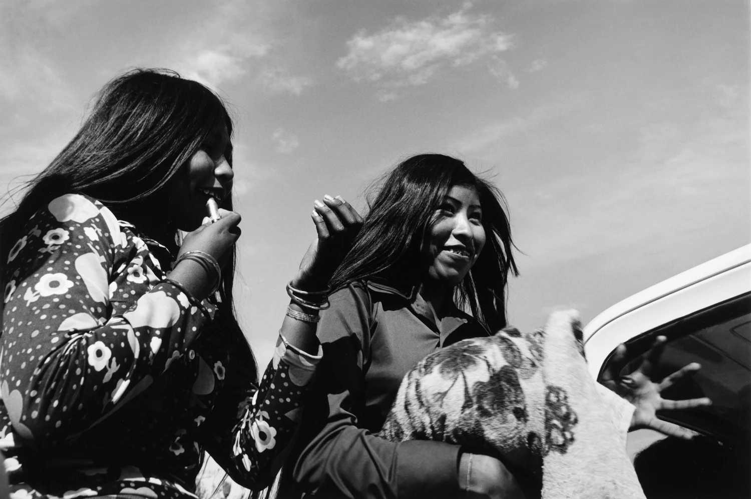 Black-and-white photograph of two women beside a car, one holding a folded blanket patterned with a tiger’s face.