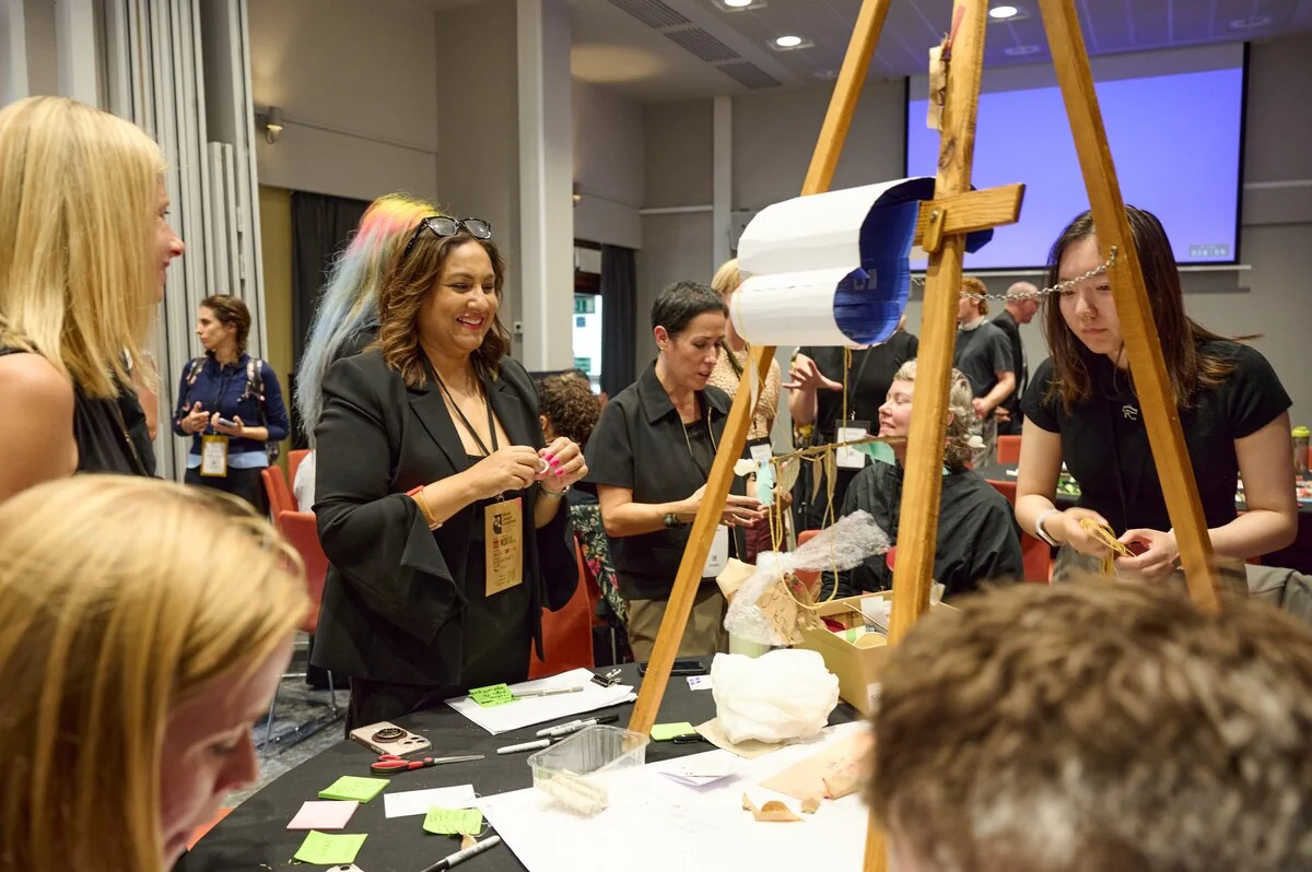 Workshop participants work with paper, string and small objects arranged on a table around a wooden easel.