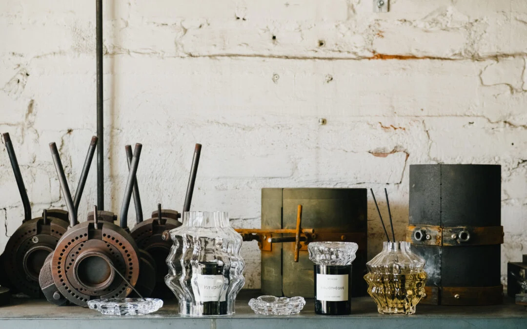 Clear sculptural glass candle and fragrance vessels stand on a workbench in front of metal glassmaking moulds and a white brick wall.